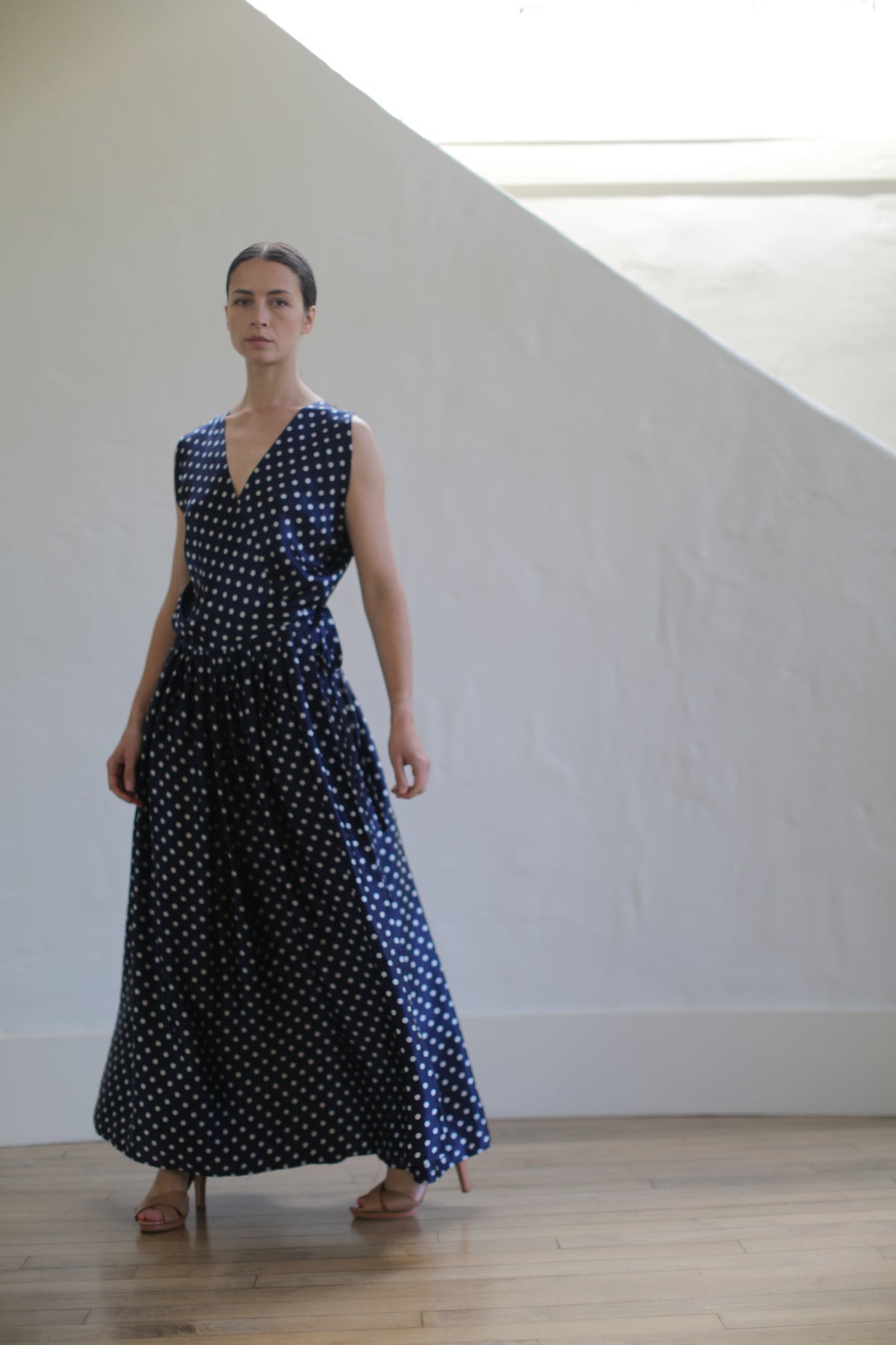 A person stands indoors against a white wall, wearing the Charmeuse Wrap Dress | Navy Polkadot and brown sandals. The dress features Japanese silk grosgrain straps, adding a touch of elegance. They are posed with one hand relaxed by their side and the other holding the side of the dress.