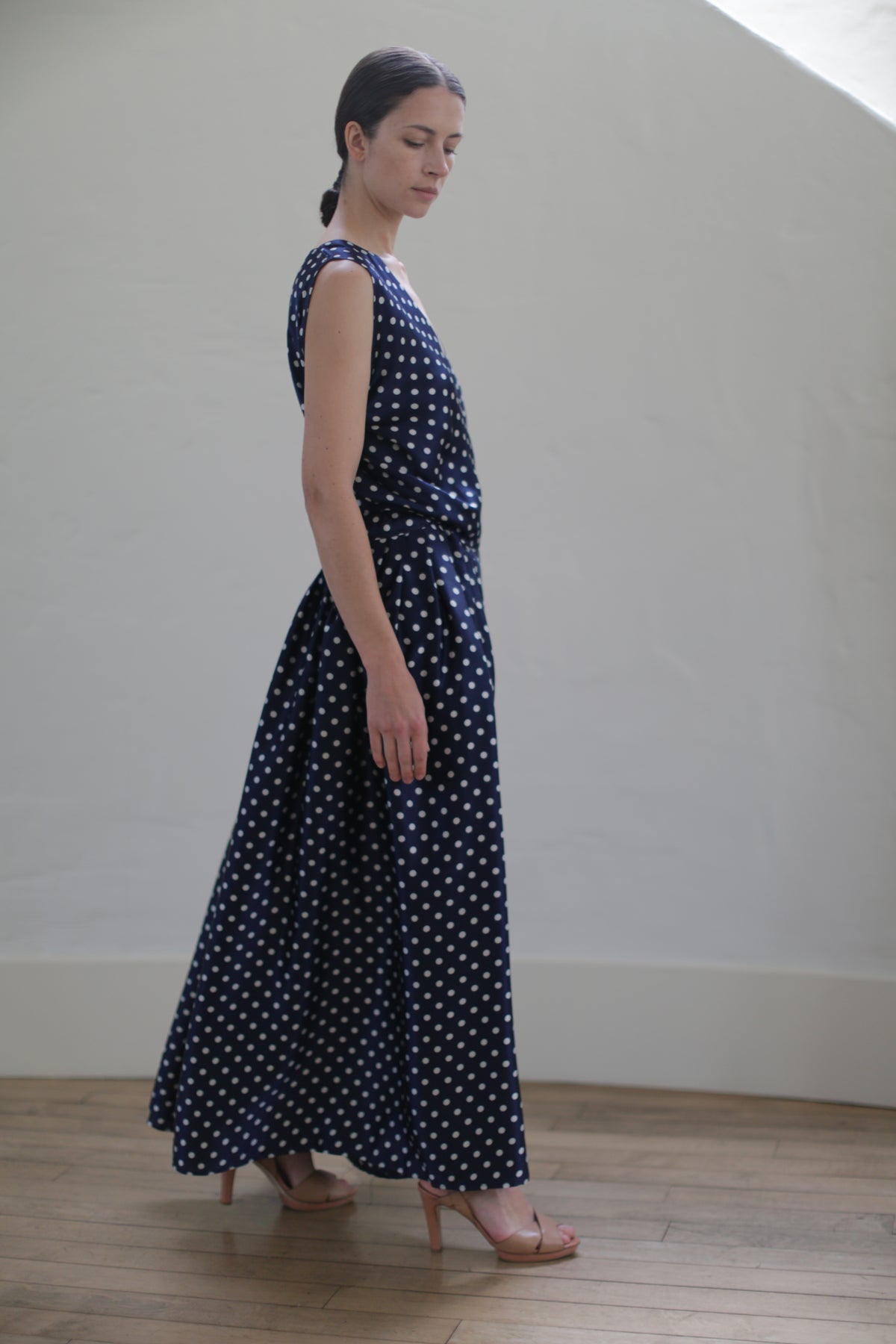 A woman stands in a minimalist room wearing a Charmeuse Wrap Dress | Navy Polkadot. She has her hair pulled back and is wearing nude-colored high heels. She is looking down and slightly away, with a soft light illuminating the scene.