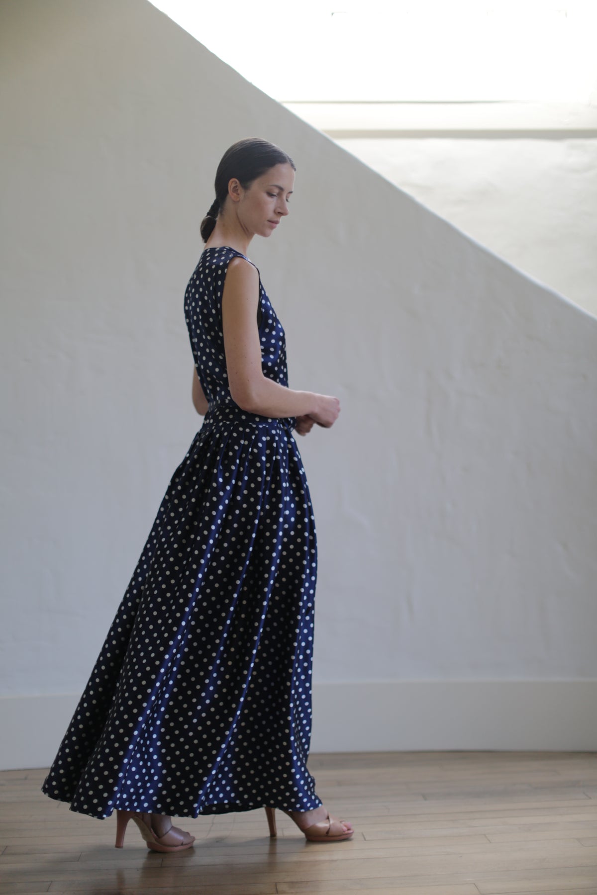A woman in a Charmeuse Wrap Dress | Navy Polkadot, accented with Japanese silk grosgrain straps, stands in profile, slightly looking down and towards her back. She has her hair tied back in a low ponytail and wears beige high heels. The background features a white wall with a large, light-filled window.
