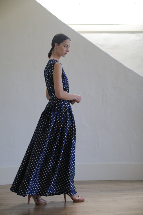 A woman in a Charmeuse Wrap Dress | Navy Polkadot, accented with Japanese silk grosgrain straps, stands in profile, slightly looking down and towards her back. She has her hair tied back in a low ponytail and wears beige high heels. The background features a white wall with a large, light-filled window.