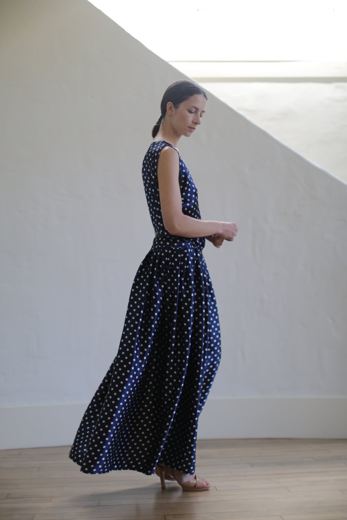 A woman in a sleeveless navy blue Charmeuse Wrap Dress | Navy Polkadot stands indoors, against a white wall. She appears to be mid-step, creating movement in the long, flowing dress. The room has a minimalistic design, with natural light illuminating the scene.