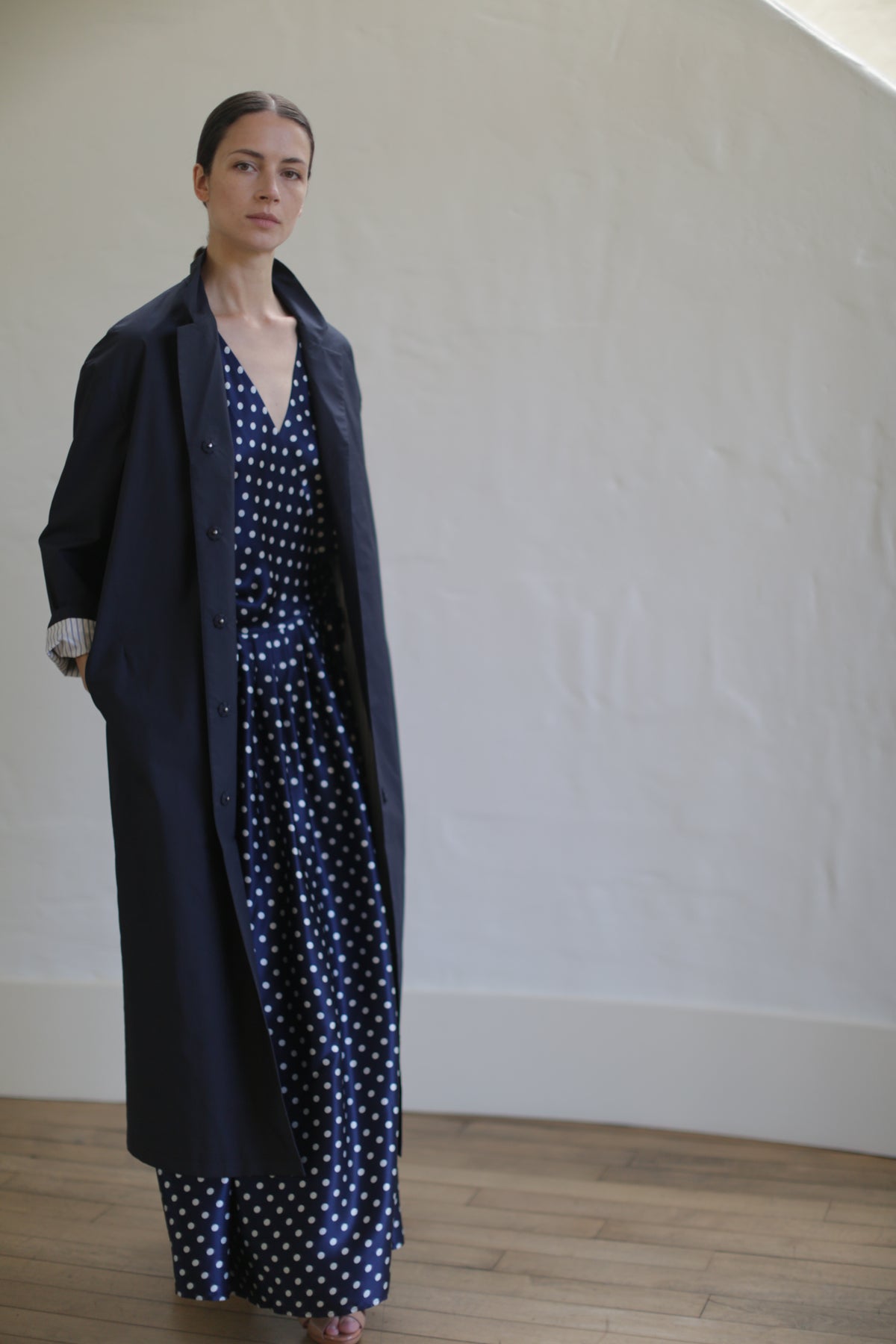 A person stands indoors against a white wall, wearing a long, dark coat over a Charmeuse Wrap Dress | Navy Polkadot. The dress features elegant Japanese silk grosgrain straps. The person looks directly at the camera with a neutral expression and has their hair pulled back. The floor is wooden and the overall setting is minimalistic.