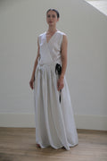 A person stands indoors against a plain white wall, wearing a long Charmeuse Wrap Dress | W/B Polkadot with black polka dots and a black ribbon accent on the waist. Handmade in San Francisco, the elegant garment complements their neutral expression and neatly pulled-back hair. The wooden floor is softly lit.