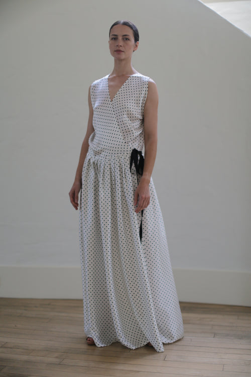 A person stands indoors against a plain white wall, wearing a long Charmeuse Wrap Dress | W/B Polkadot with black polka dots and a black ribbon accent on the waist. Handmade in San Francisco, the elegant garment complements their neutral expression and neatly pulled-back hair. The wooden floor is softly lit.