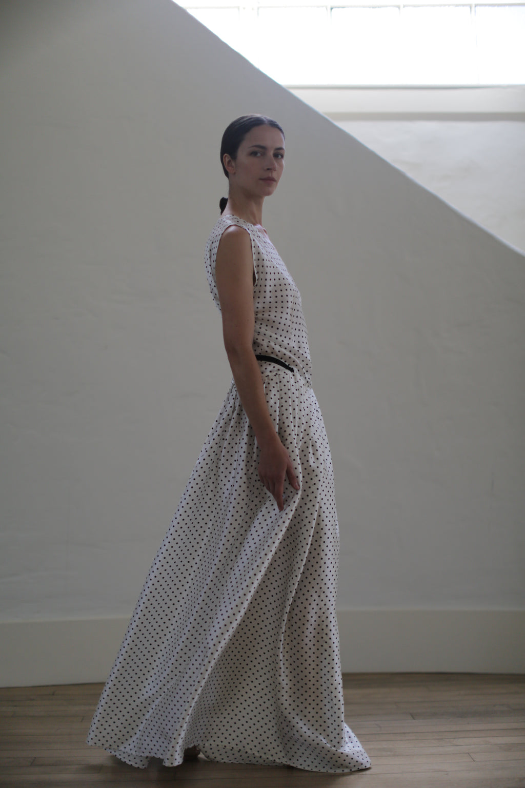A woman stands in a side pose wearing the Charmeuse Wrap Dress | W/B Polkadot. Handmade in San Francisco, the zero-waste design accentuates her pulled-back hair as she stands in a minimalist room with a large, angled window that lets soft light create a serene ambiance.