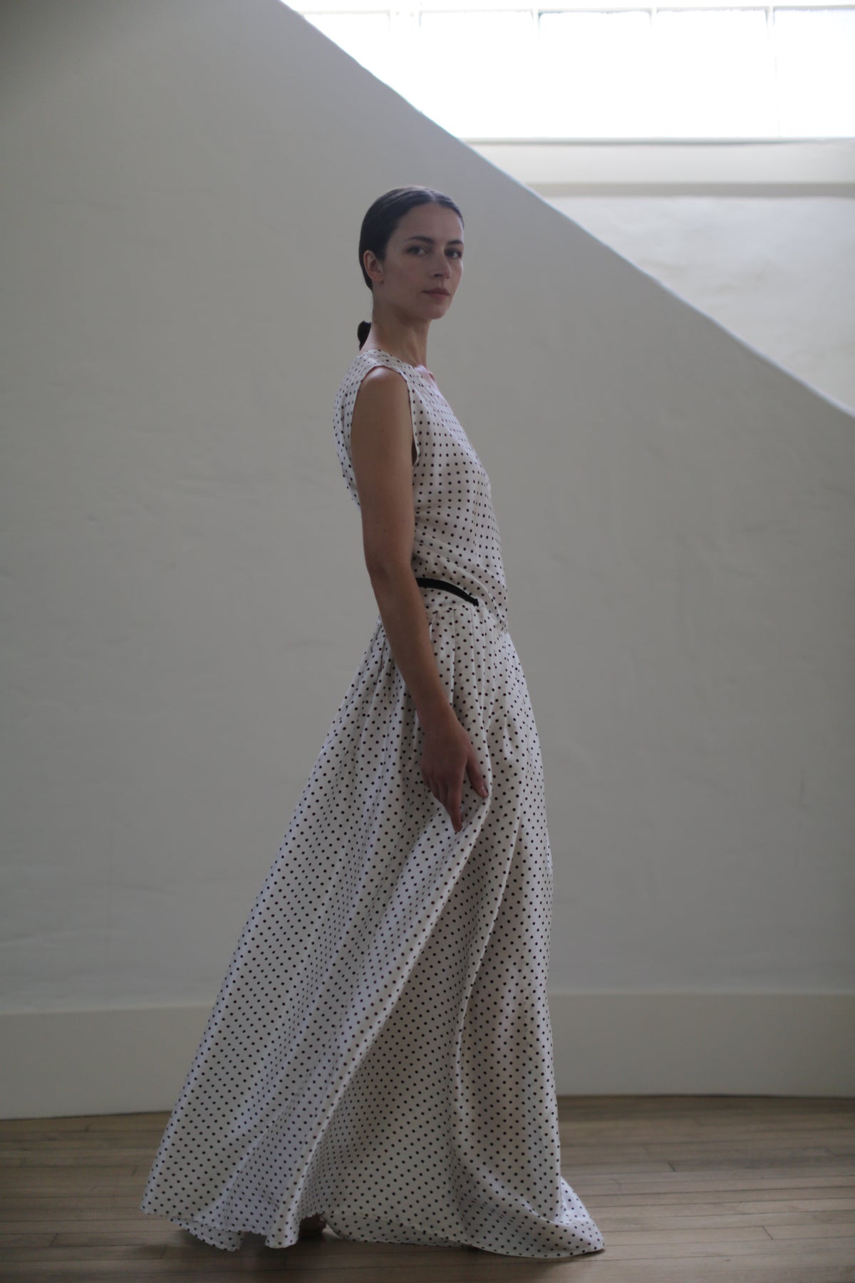 A woman stands in a side pose wearing the Charmeuse Wrap Dress | W/B Polkadot. Handmade in San Francisco, the zero-waste design accentuates her pulled-back hair as she stands in a minimalist room with a large, angled window that lets soft light create a serene ambiance.