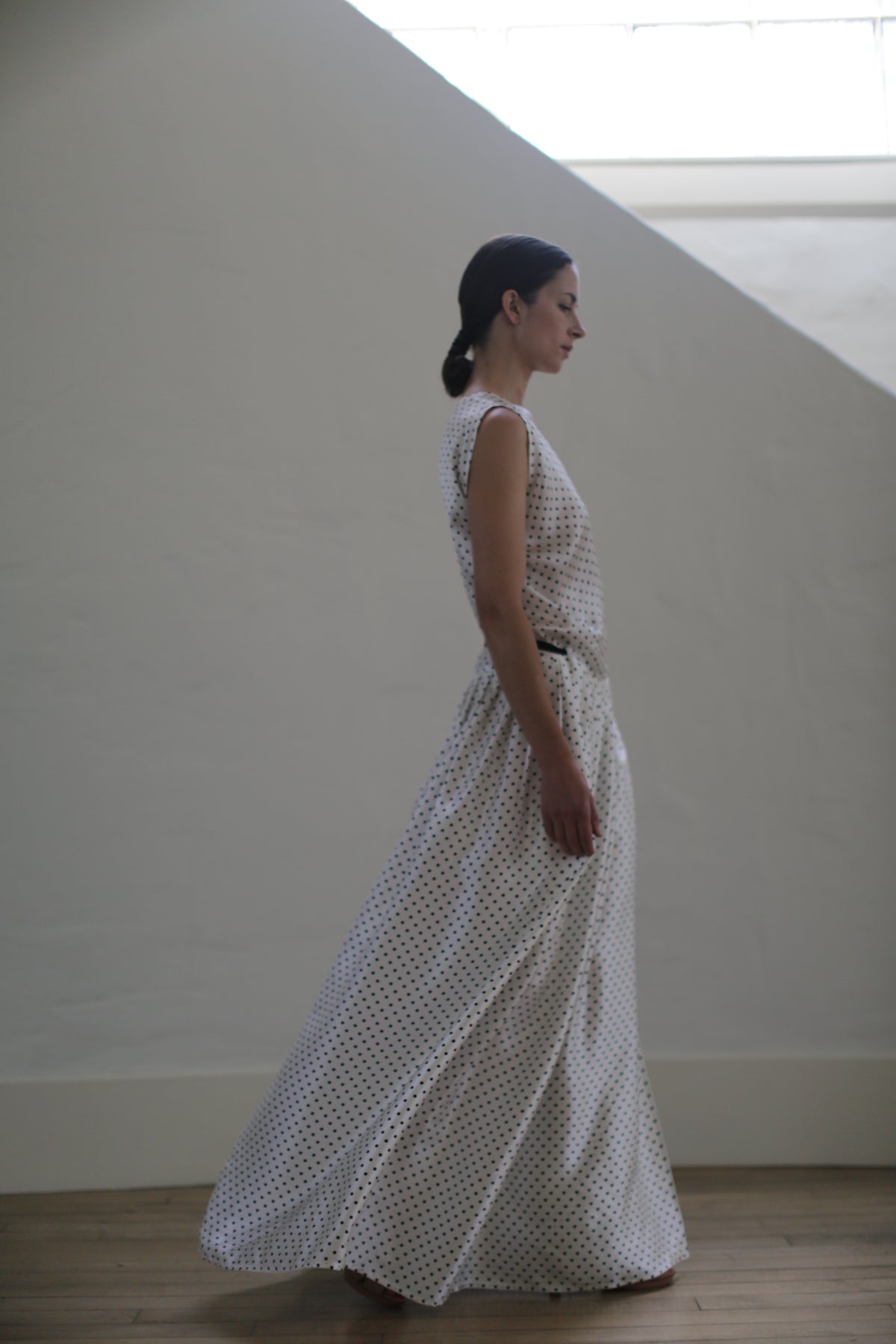 A woman in profile wearing a flowing, sleeveless Charmeuse Wrap Dress | W/B Polkadot. Her hair is pulled back into a low bun. She stands indoors against a plain, light-colored wall and wooden floor, with a large window above, casting natural light. This zero-waste design is handmade in San Francisco.