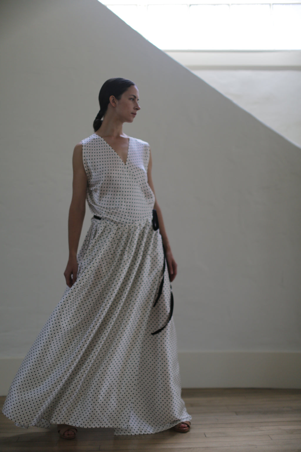 A woman stands in a softly lit room wearing a white, sleeveless Charmeuse Wrap Dress | W/B Polkadot with a flowing skirt. She is looking to her left, her hair pulled back in a ponytail. The dress, handmade in San Francisco with zero-waste design principles, complements the serene setting of the white wall and wooden floor.