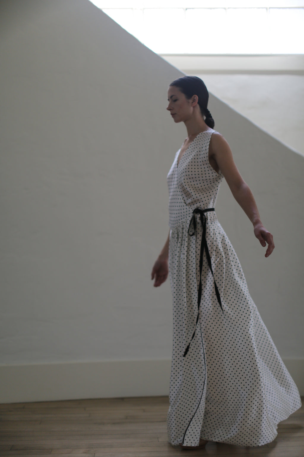 A woman in profile is standing indoors against a minimalist, light-colored wall. She is wearing a Charmeuse Wrap Dress | W/B Polkadot. Handmade in San Francisco, her attire complements her serene, poised demeanor as her hair is pulled back into a low ponytail.