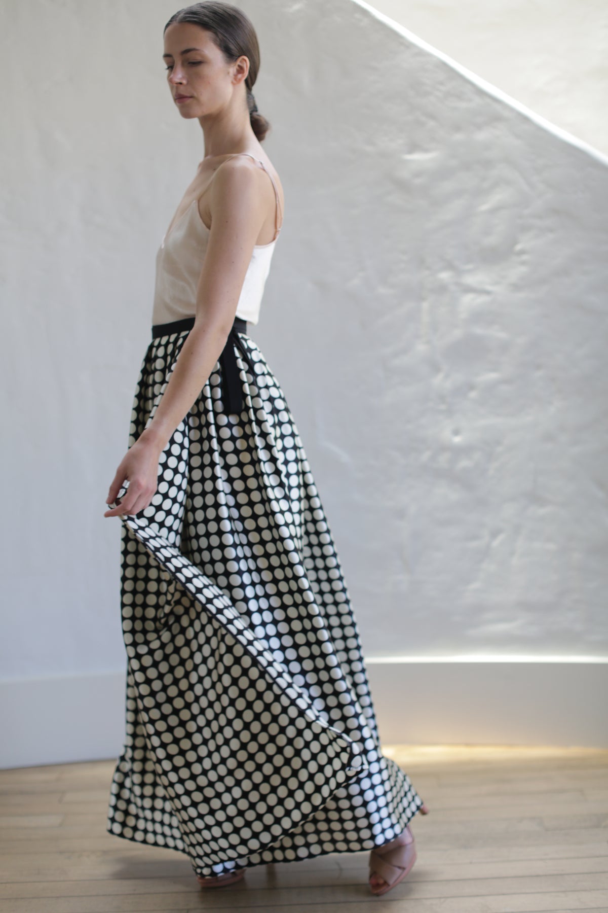 A person with long dark hair tied back is wearing a sleeveless beige top and a Charmeuse Pleated Long Wrap Skirt | Big Polkadot. They are standing in profile, slightly turning, and holding the skirt with one hand. The background features a textured white wall and wooden flooring.