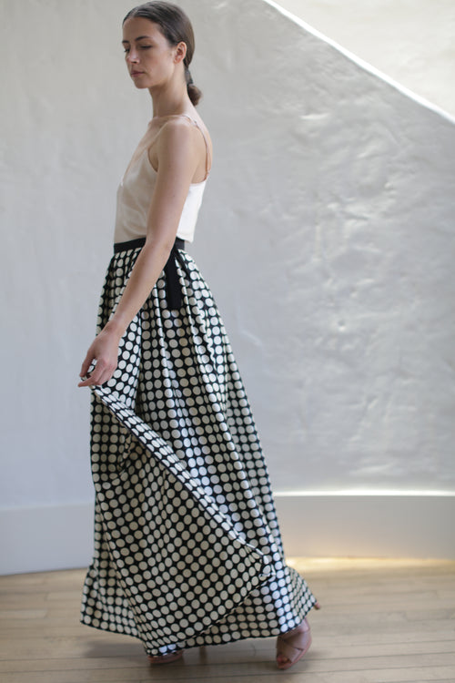 A person with long dark hair tied back is wearing a sleeveless beige top and a Charmeuse Pleated Long Wrap Skirt | Big Polkadot. They are standing in profile, slightly turning, and holding the skirt with one hand. The background features a textured white wall and wooden flooring.
