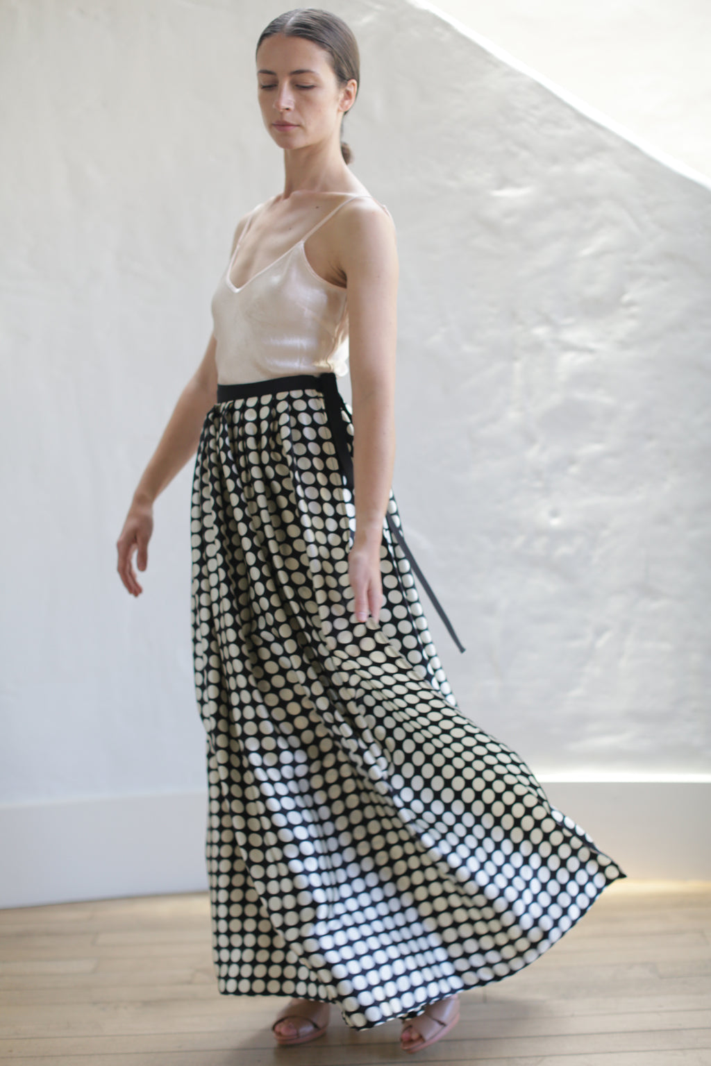 A woman with long hair, wearing a sleeveless beige top and a Charmeuse Pleated Long Wrap Skirt | Big Polkadot, stands with one arm slightly extended. She is looking down and appears to be in motion, with a white textured wall and light wooden floor in the background.