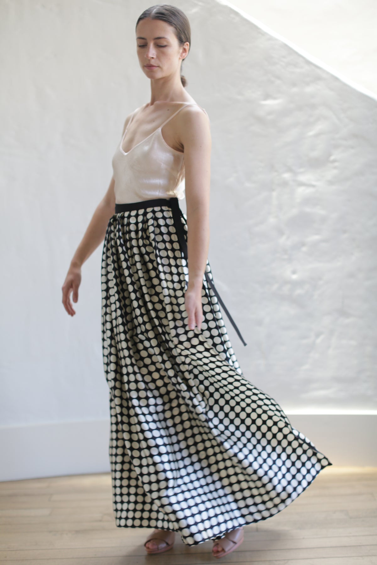 A woman with long hair, wearing a sleeveless beige top and a Charmeuse Pleated Long Wrap Skirt | Big Polkadot, stands with one arm slightly extended. She is looking down and appears to be in motion, with a white textured wall and light wooden floor in the background.