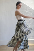A woman with dark hair is gracefully twirling, wearing a sleeveless white top and a Charmeuse Pleated Long Wrap Skirt | Big Polkadot. She is barefoot on a wooden floor, and the background is a minimalist white wall with natural light illuminating the scene.