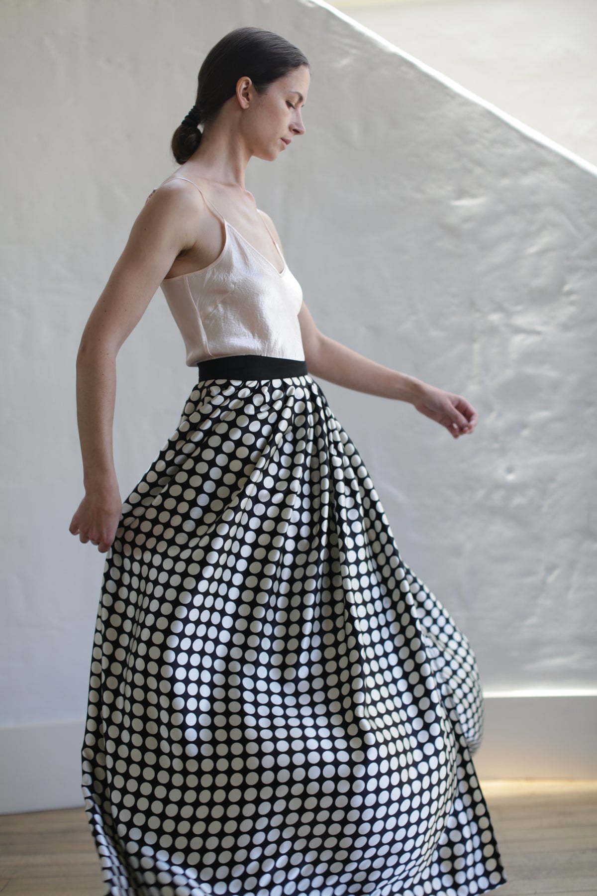 A person with dark hair in a low bun is wearing a light-colored sleeveless top and a Charmeuse Pleated Long Wrap Skirt | Big Polkadot. The charmeuse silk fabric catches the light as they pose sideways, slightly looking down, in a minimalist, light-filled room with a textured white wall.