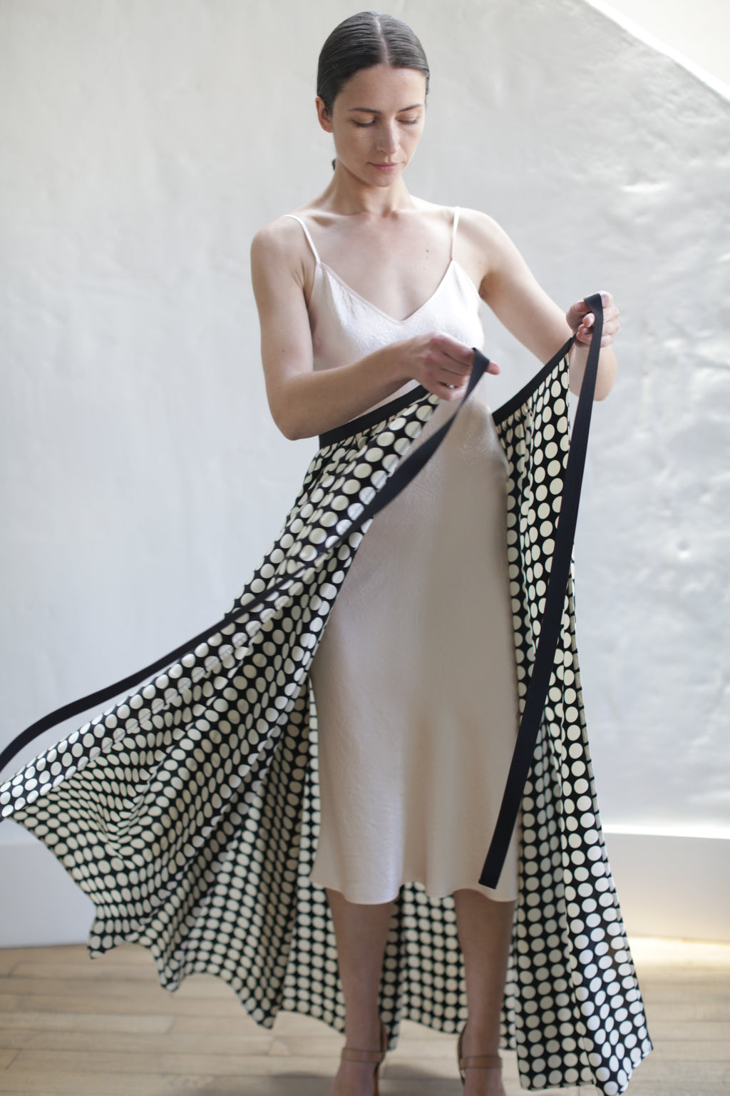 A woman in a light pink slip dress stands in a room with a white wall, holding and displaying a Charmeuse Pleated Long Wrap Skirt | Big Polkadot. She appears to be spreading the fabric outwards with both hands, and her hair is pulled back.