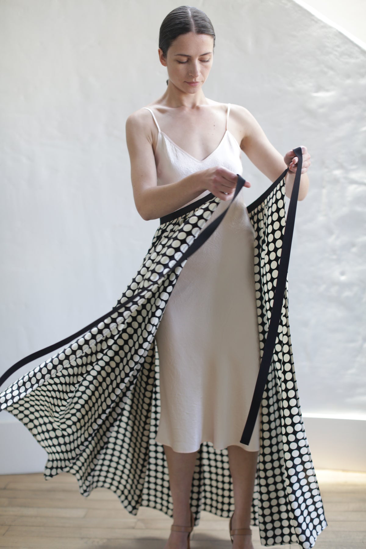 A woman in a light pink slip dress stands in a room with a white wall, holding and displaying a Charmeuse Pleated Long Wrap Skirt | Big Polkadot. She appears to be spreading the fabric outwards with both hands, and her hair is pulled back.