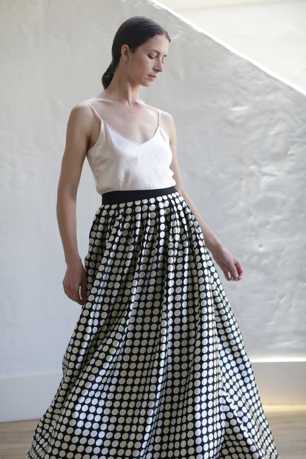 A woman in a white spaghetti strap top and a Charmeuse Pleated Long Wrap Skirt | Big Polkadot. The charmeuse silk fabric catches the light beautifully as she stands in front of a white, textured wall, looking down and to her left.