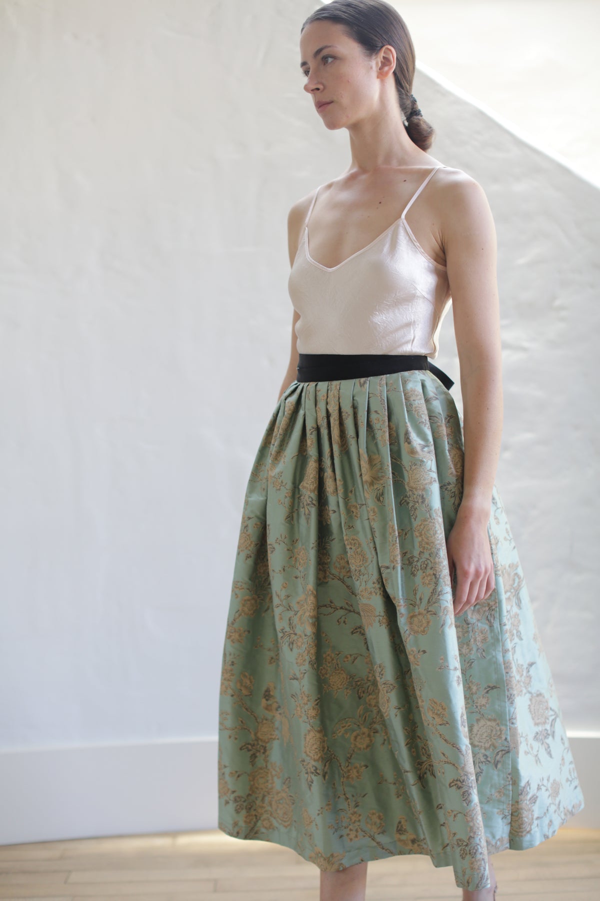 A woman stands against a light-colored textured wall, wearing a limited edition light pink spaghetti strap top and a Dupioni Pleated Wrap Skirt | Jade. Her hair is pulled back, and she gazes slightly to the side. Light streams in from the right.