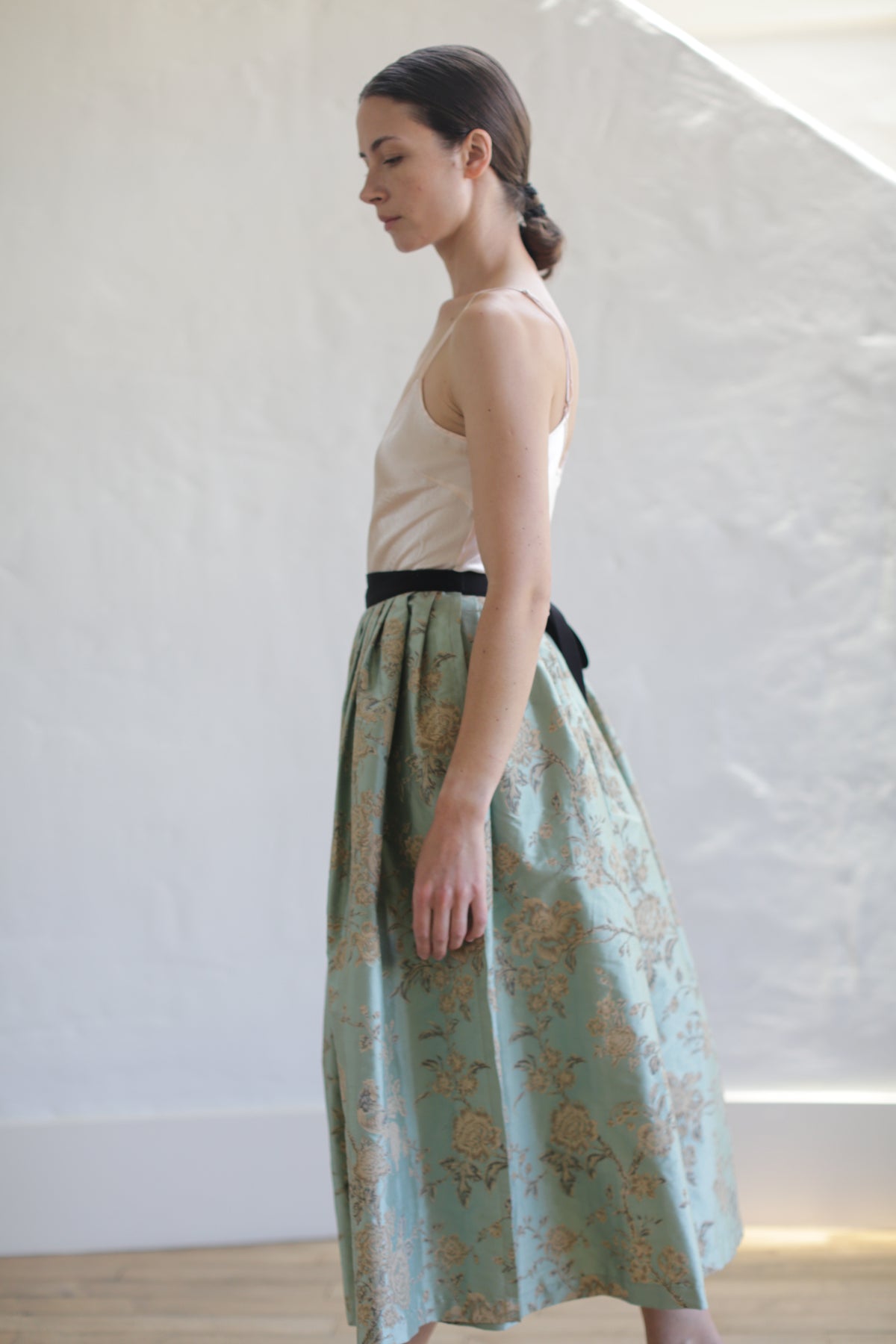 A woman with her hair pulled back wears a light beige, handmade sleeveless top and a long, high-waisted skirt crafted from limited edition Dupioni Pleated Wrap Skirt | Jade featuring a floral pattern in shades of green and gold. She stands in profile against a neutral, textured background.