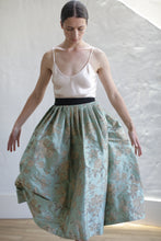 Load image into Gallery viewer, A woman gracefully poses indoors, wearing a light pink spaghetti strap top and a high-waisted, full, floral-patterned blue-green skirt with gold accents. Her hair is pulled back, and she stands against a textured white wall on a wooden floor. This stunning outfit is crafted from limited edition Dupioni Pleated Wrap Skirt | Jade.