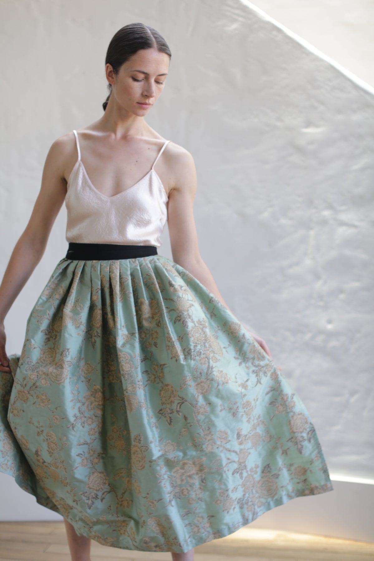 A woman stands gracefully, wearing a white spaghetti-strap top and a Dupioni Pleated Wrap Skirt | Jade. The environment is softly lit, and she has her eyes closed, looking serene. The background features a textured, white wall with angled lighting.