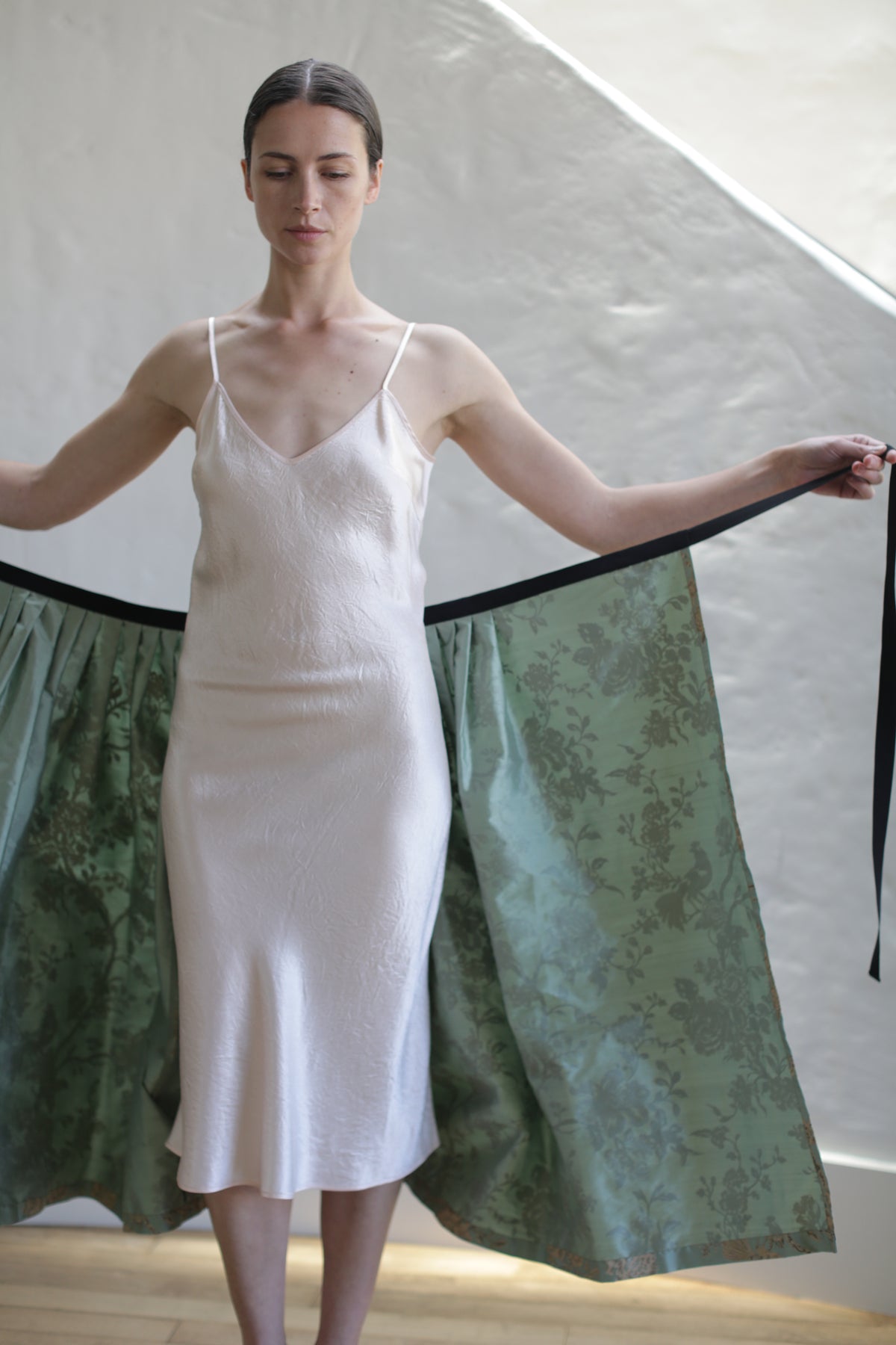 A woman in a white, sleeveless, knee-length dress stands indoors with her eyes closed. She holds the sides of a long, green, floral-patterned Dupioni Pleated Wrap Skirt | Jade that drapes from her shoulders. The backdrop is a plain, off-white wall partially lit by natural light.