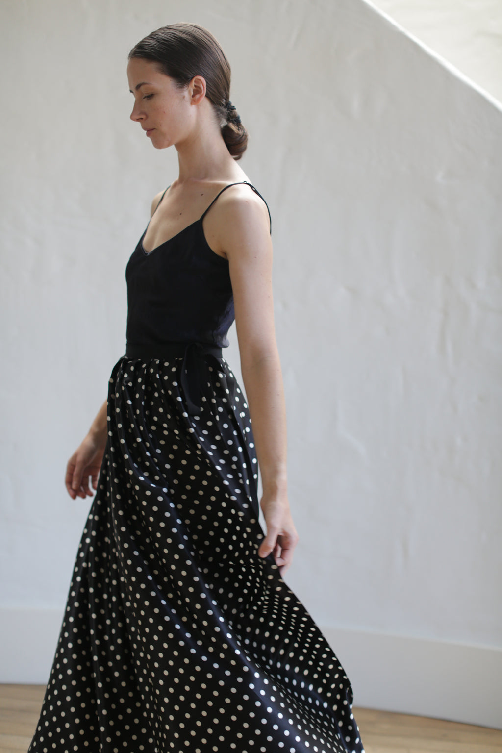 A woman wearing a sleeveless black top and a Charmeuse Pleated Long Wrap Skirt | B/W Polkadot is seen standing in profile. Her dark hair is tied back in a low bun. The background is a plain white wall and wooden floor.