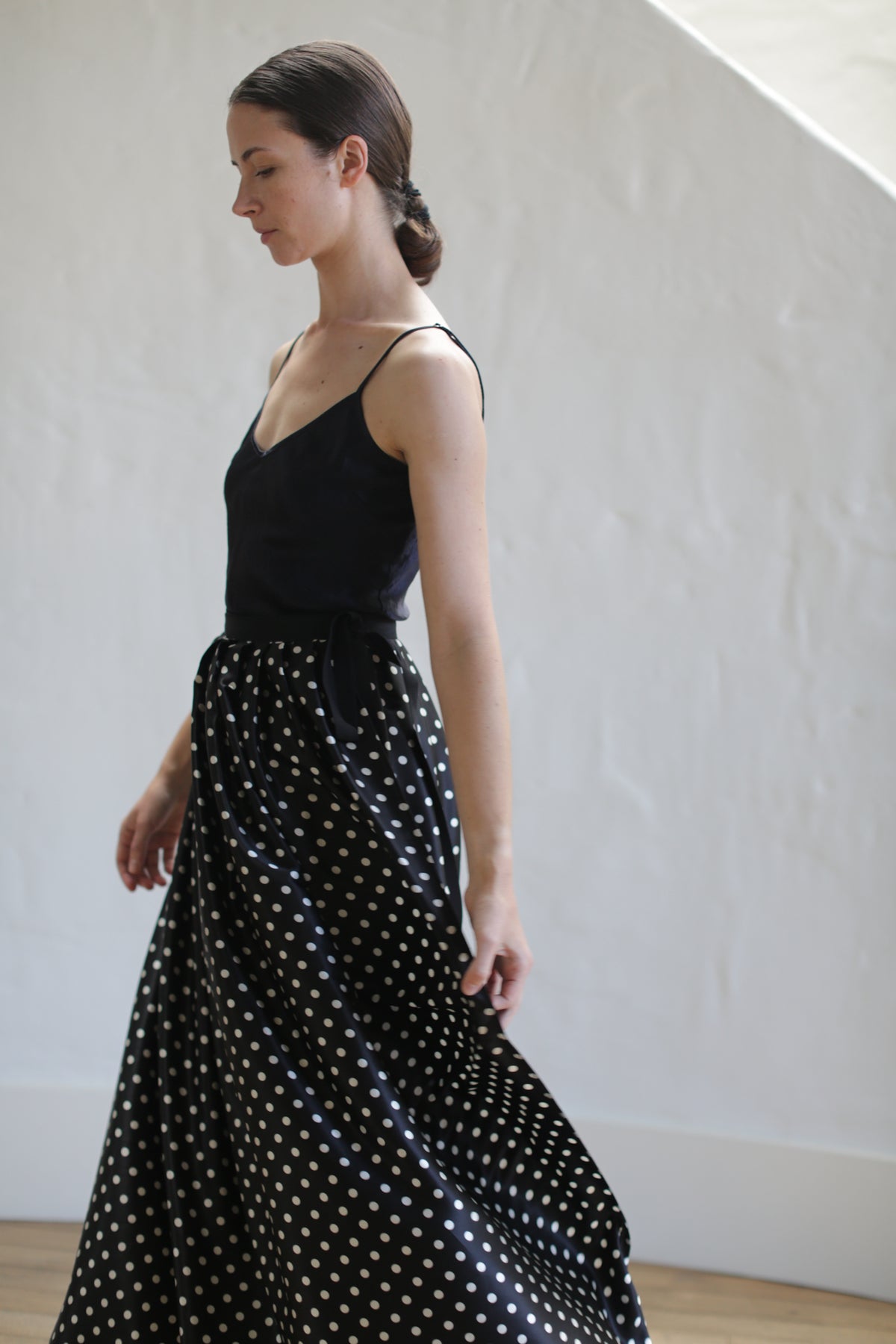 A woman wearing a sleeveless black top and a Charmeuse Pleated Long Wrap Skirt | B/W Polkadot is seen standing in profile. Her dark hair is tied back in a low bun. The background is a plain white wall and wooden floor.