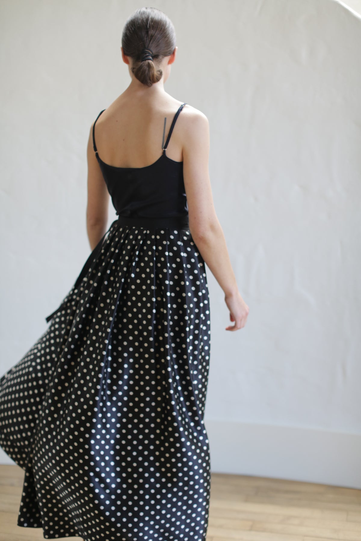 A woman with her hair in a low bun, wearing a sleeveless black top and a Charmeuse Pleated Long Wrap Skirt | B/W Polkadot, is photographed from behind. She is standing near a light-colored wall on a wooden floor.
