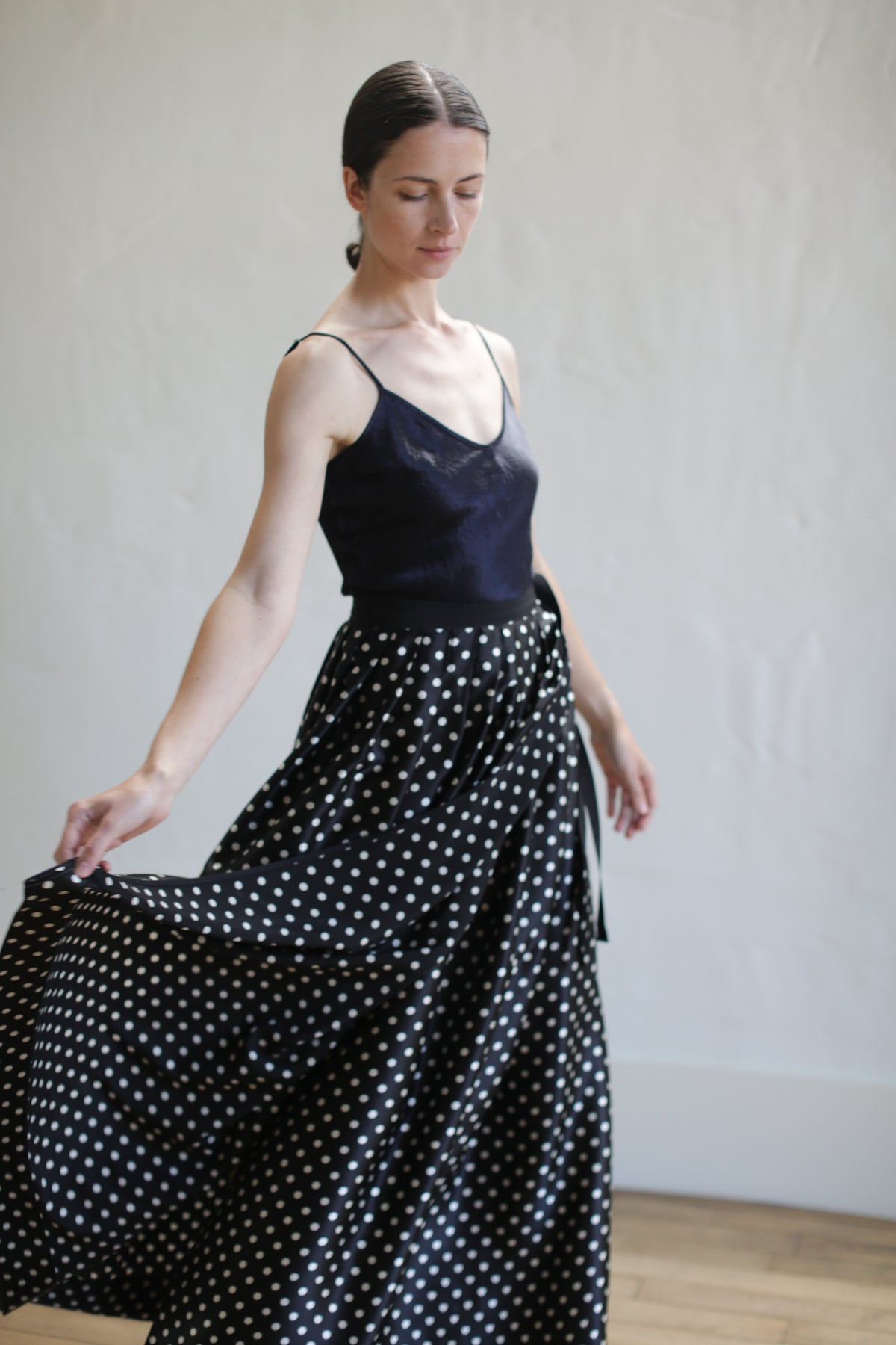 A woman in a sleeveless, black top and a flowing Charmeuse Pleated Long Wrap Skirt | B/W Polkadot stands against a light-colored wall. She holds part of her wrap skirt with one hand while looking down, creating a graceful and serene pose.