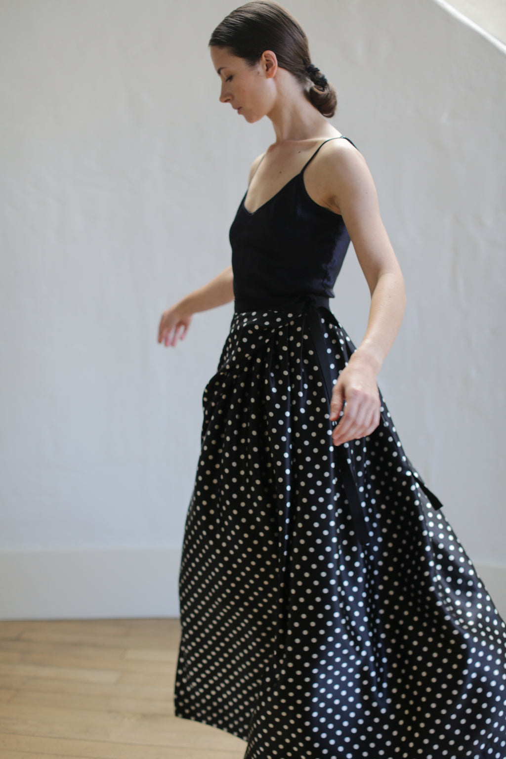A woman with dark hair in a low bun is wearing a sleeveless black top and a Charmeuse Pleated Long Wrap Skirt | B/W Polkadot. She is captured mid-motion, looking to her left, with a plain white wall in the background.