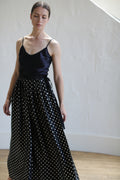 A person with closed eyes is wearing a black charmeuse silk sleeveless top and a Charmeuse Pleated Long Wrap Skirt | B/W Polkadot. They are standing against a light-colored, textured wall with wooden flooring beneath them. The light creates subtle shadows on the wall.