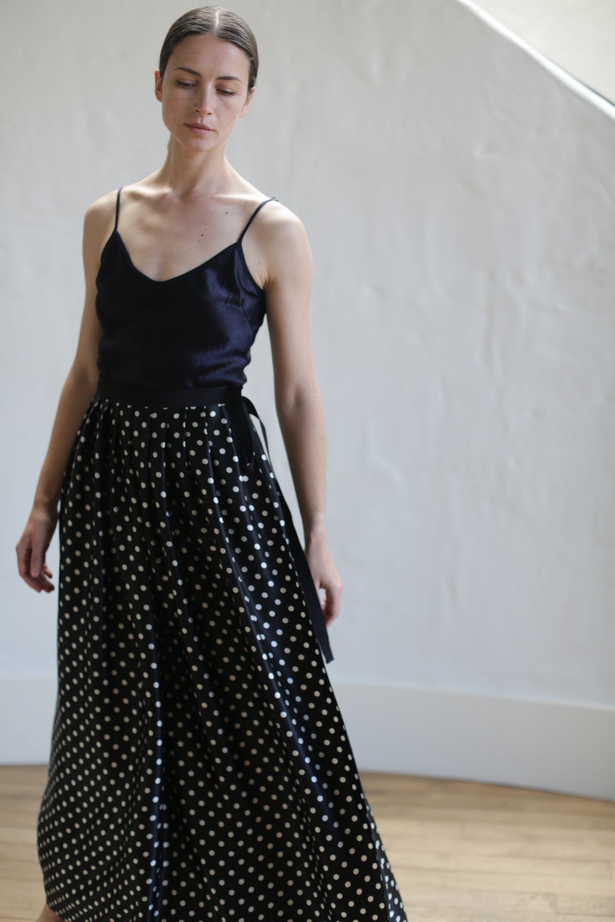 A person with closed eyes is wearing a black charmeuse silk sleeveless top and a Charmeuse Pleated Long Wrap Skirt | B/W Polkadot. They are standing against a light-colored, textured wall with wooden flooring beneath them. The light creates subtle shadows on the wall.