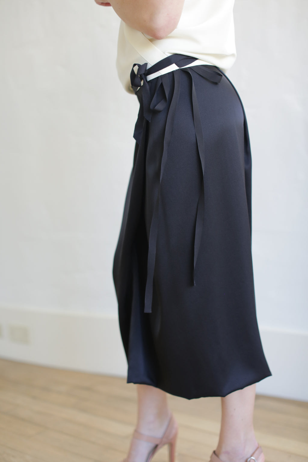 A person in a Charmeuse Petal Wrap Skirt in black, paired with a white top and beige high-heeled sandals, stands on a wooden floor against a light backdrop. The zero-waste design skirt features long ties at the waist.