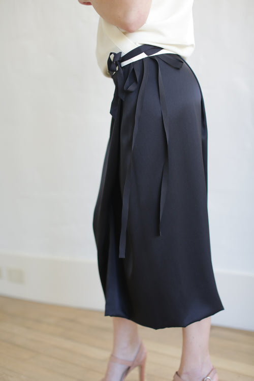 A person in a Charmeuse Petal Wrap Skirt in black, paired with a white top and beige high-heeled sandals, stands on a wooden floor against a light backdrop. The zero-waste design skirt features long ties at the waist.