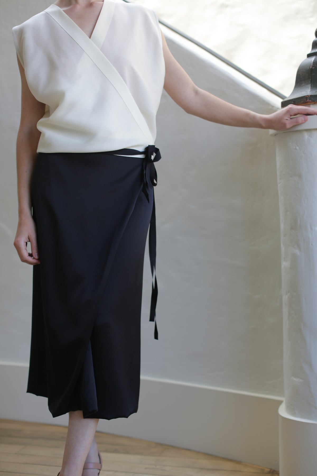 A person in a sleeveless white blouse and the elegant Charmeuse Petal Wrap Skirt in black, featuring a cashmere waistband and zero-waste design, stands by a curved white railing with one hand resting on it. The softly lit, neutral background enhances the outfit's simple elegance.