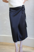 A person in a white top wears the Charmeuse Petal Wrap Skirt in black, which features a tied bow on the side. The neutral background includes a light-colored wall and wooden floor, and their feet are in light-hued open-toe shoes.