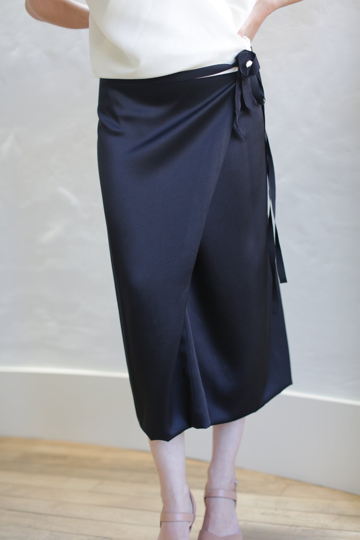 A person in a white top wears the Charmeuse Petal Wrap Skirt in black, which features a tied bow on the side. The neutral background includes a light-colored wall and wooden floor, and their feet are in light-hued open-toe shoes.
