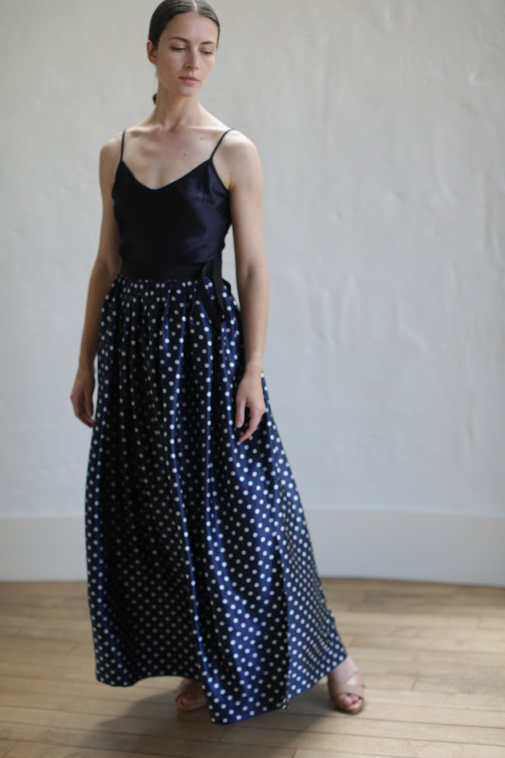 A person stands on a wooden floor in front of a white wall, wearing a black sleeveless top and a Charmeuse Pleated Long Wrap Skirt | Navy Polkadot. Their head is slightly turned to one side, and their eyes are closed.