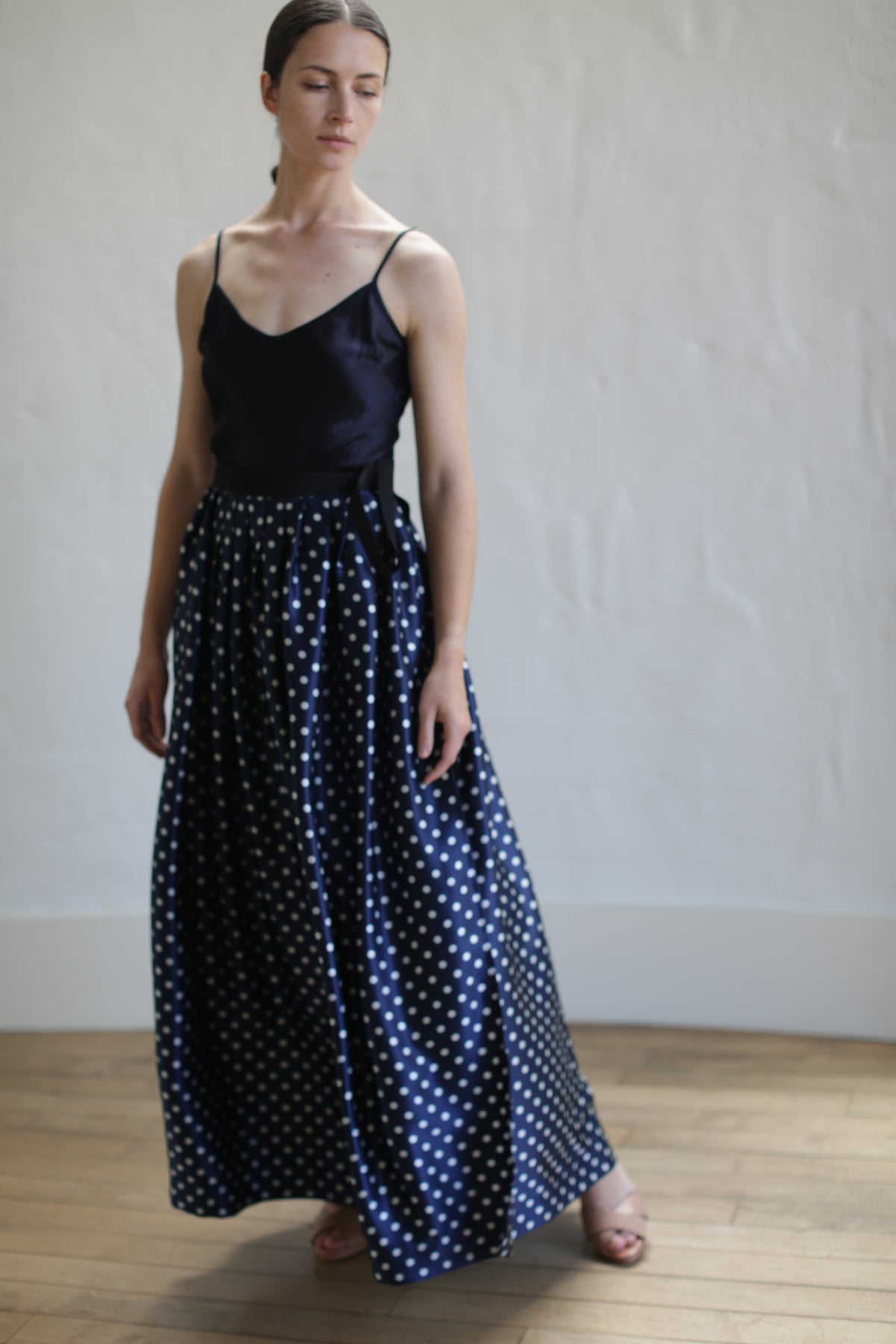 A person stands on a wooden floor in front of a white wall, wearing a black sleeveless top and a Charmeuse Pleated Long Wrap Skirt | Navy Polkadot. Their head is slightly turned to one side, and their eyes are closed.