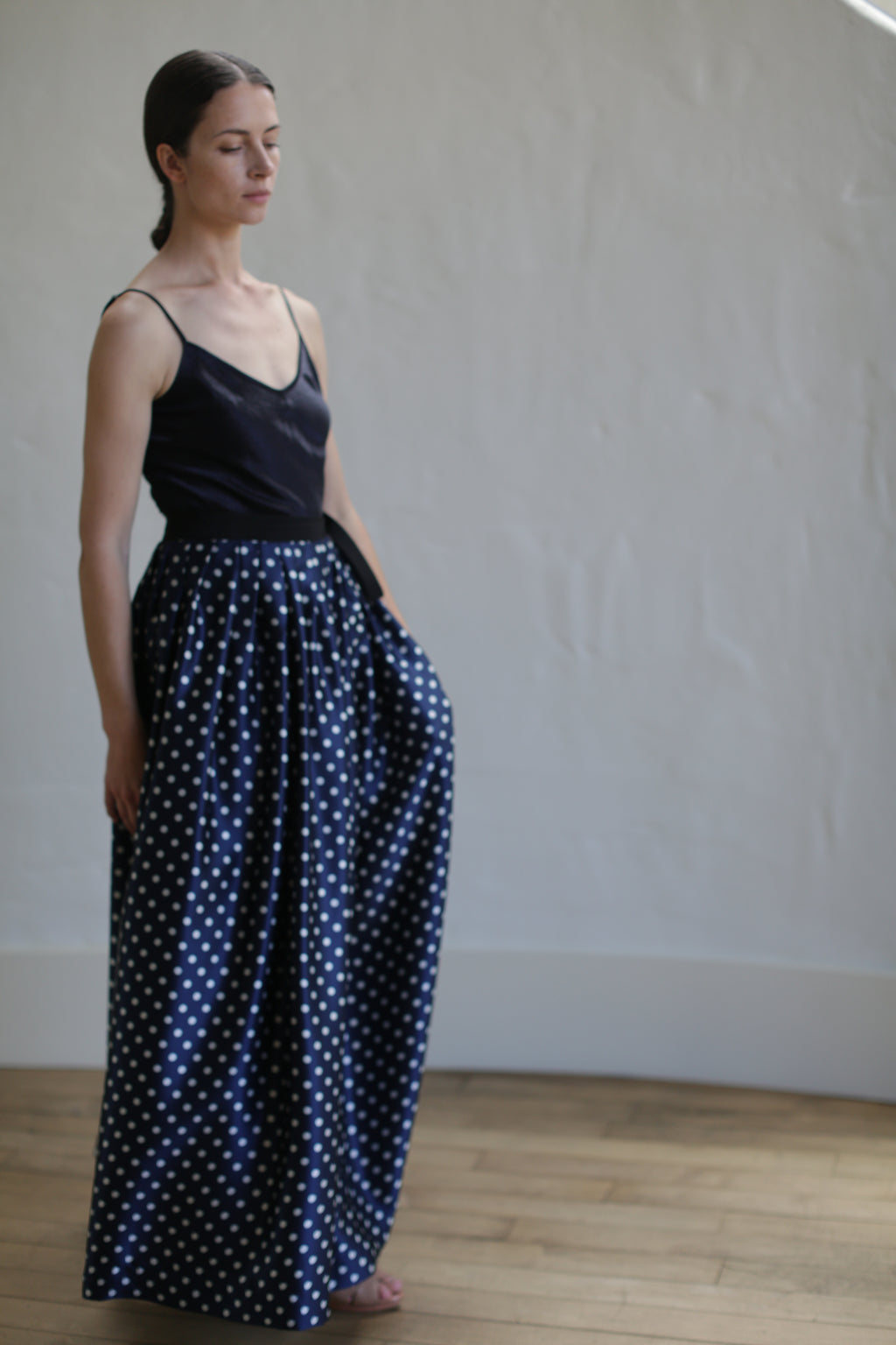 A person with long hair, wearing a black spaghetti-strap top and a high-waisted, **Charmeuse Pleated Long Wrap Skirt | Navy Polkadot**, stands indoors against a plain light-colored wall. Their eyes are closed, and one hand is in the skirt pocket.