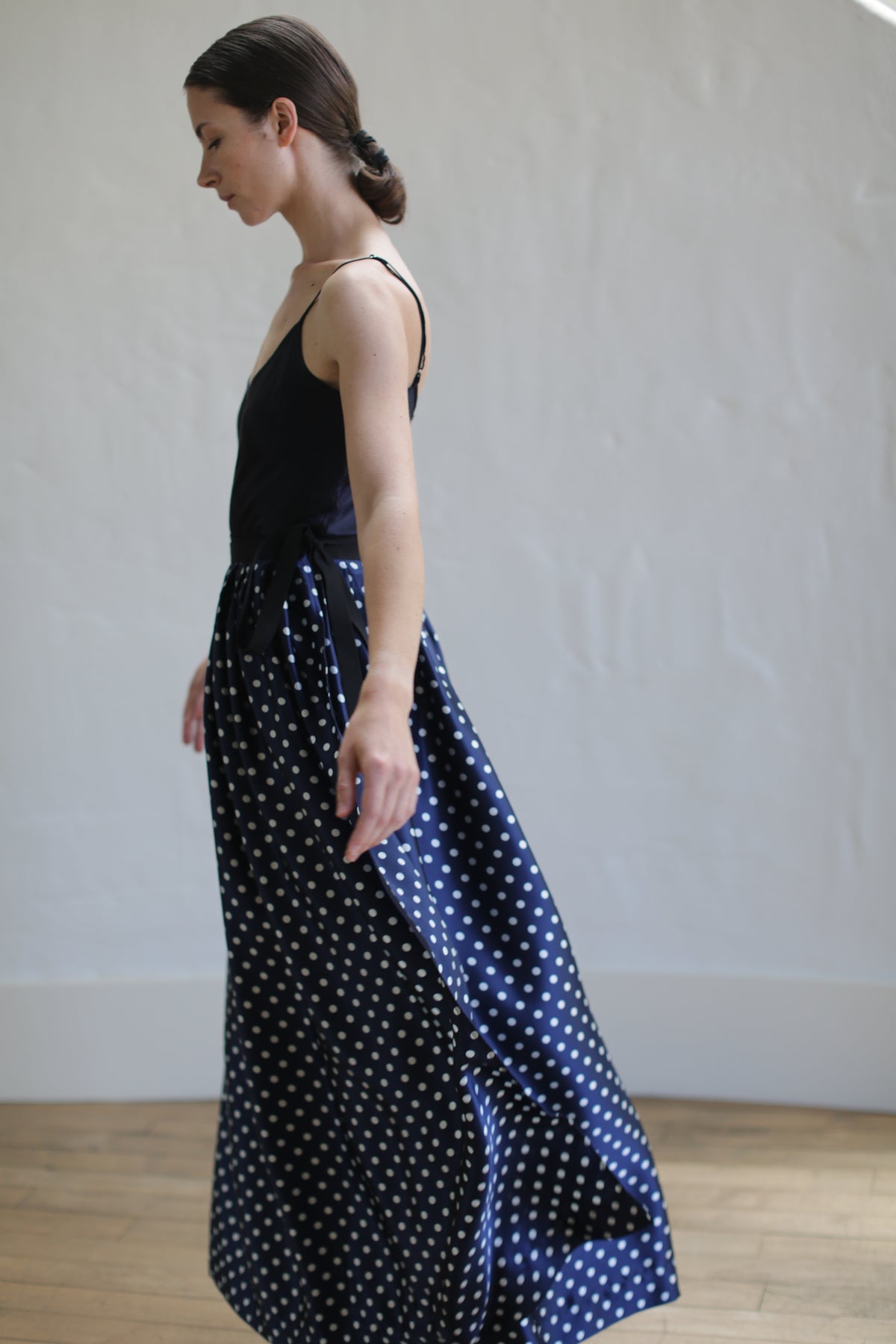 A woman with dark hair tied back in a low bun is standing in profile, wearing a black sleeveless top and a Charmeuse Pleated Long Wrap Skirt | Navy Polkadot. She is in a well-lit room with light wood flooring and a plain white wall in the background.