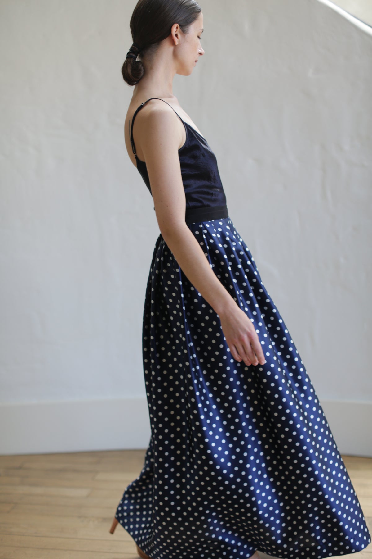 A woman in profile wearing a sleeveless black top and a Charmeuse Pleated Long Wrap Skirt | Navy Polkadot stands in a room with a light-colored wooden floor and a white wall. Her dark hair is neatly tied back in a low bun.