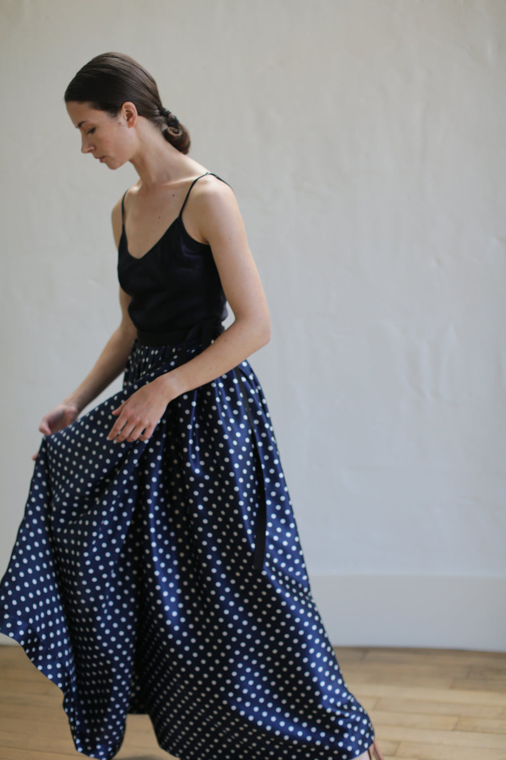 A woman with dark hair tied back is wearing a black sleeveless top and a Charmeuse Pleated Long Wrap Skirt | Navy Polkadot. She is captured in profile, with one hand holding the wrap skirt as if in motion. The background is a plain, light-colored wall.