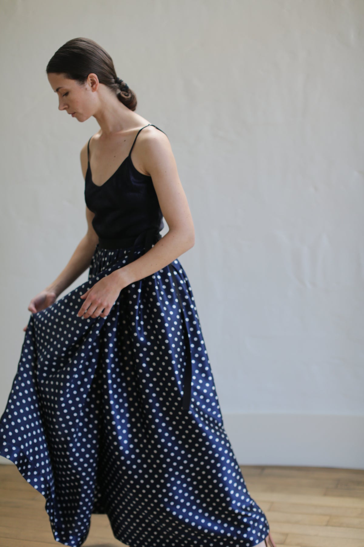A woman with dark hair tied back is wearing a black sleeveless top and a Charmeuse Pleated Long Wrap Skirt | Navy Polkadot. She is captured in profile, with one hand holding the wrap skirt as if in motion. The background is a plain, light-colored wall.