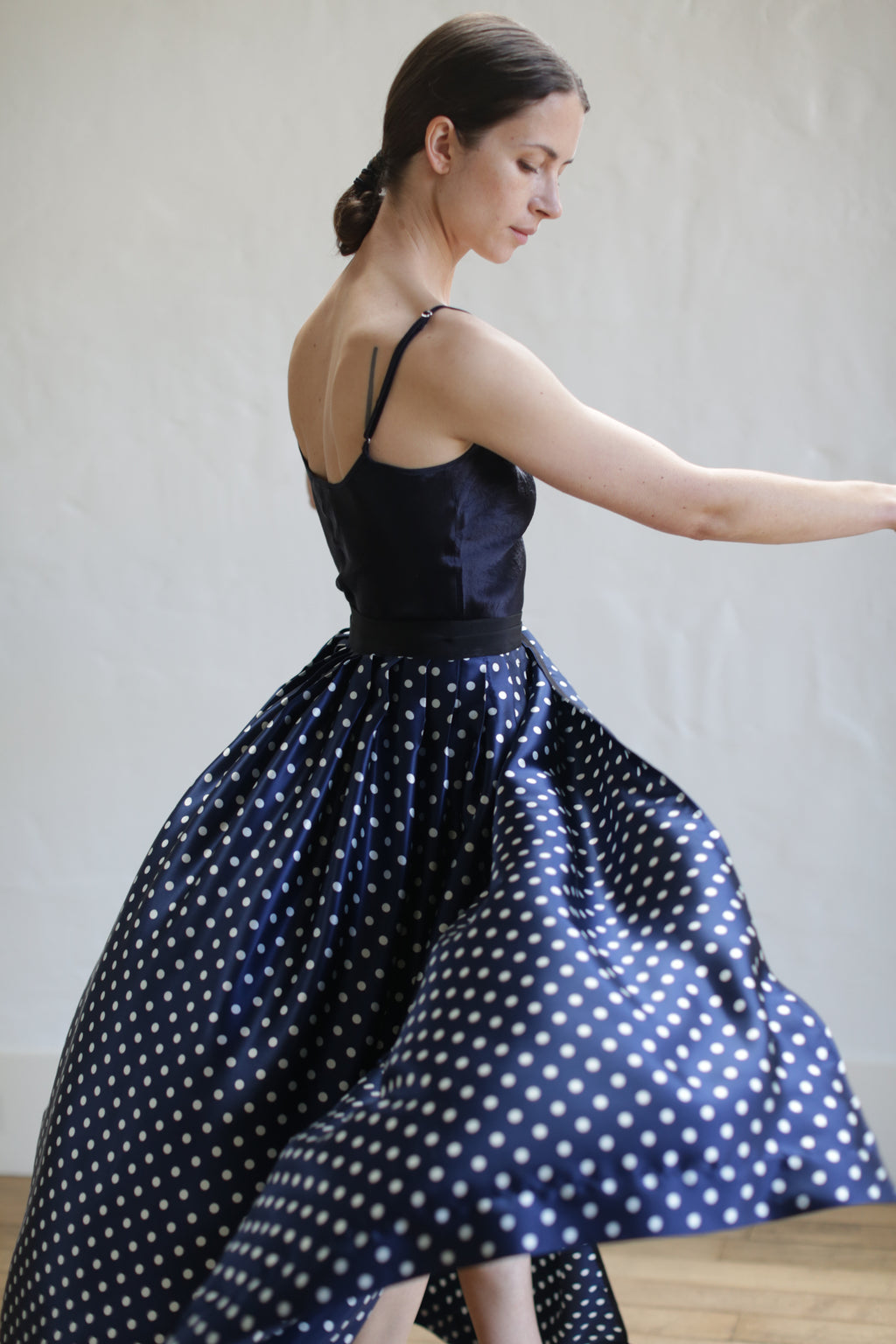 A woman with dark hair styled in a low bun is twirling in a sleeveless black and white polka dot dress with a long, flowing Charmeuse Pleated Long Wrap Skirt | Navy Polkadot. She is pictured from the side against a plain, light-colored background.