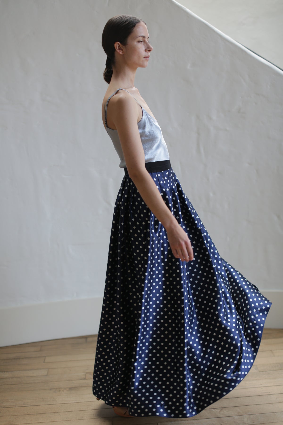 A person with long, dark hair tied back is wearing a silver satin top and a Charmeuse Pleated Long Wrap Skirt | Navy Polkadot. The person is standing indoors against a light-colored wall and wooden floor, facing slightly to the left and looking ahead.