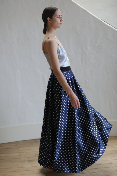 A person with long, dark hair tied back is wearing a silver satin top and a Charmeuse Pleated Long Wrap Skirt | Navy Polkadot. The person is standing indoors against a light-colored wall and wooden floor, facing slightly to the left and looking ahead.