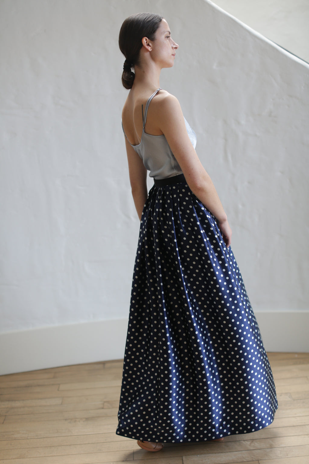 A woman stands sideways, looking forward. She is wearing a light gray sleeveless top and a Charmeuse Pleated Long Wrap Skirt | Navy Polkadot. Her hair is tied back in a low ponytail. The background consists of a white wall and wooden flooring.